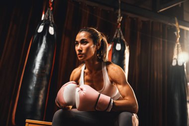 Asyalı kadın Muay Thai boksörü ya da kickboks kısa bir aradan sonra eldivenlerini giymiş ve arka planda boks ekipmanları ile salonda oturuyor. Güçlü ve kaslı vücut sporcusu. Impetus