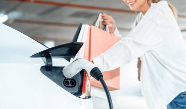 Genç bir kadın EV elektrikli arabasıyla alışveriş merkezinin otoparkına gidiyor şehir merkezinde elektrik iç organlarının yeşil temiz şarj edilebilir enerji ile sürdürülebilirlik yaşam tarzını gösteriyor.