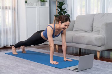 Spor kıyafetli Asyalı bir kadın antrenman rutini olarak spor minderinin üzerinde burpee yapıyor. Çekici kız sağlıklı yaşam tarzını online egzersiz videosuyla takip ediyor. Kuvvetli