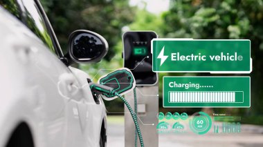 EV şarj kablosuna elektrik kablosu takılı. Çevre yeşil parkı ve yeşillik arka planında akıllı dijital batarya durum hologramını gösteriyor. Enerji sürdürülebilirliği teknolojik gelişim.