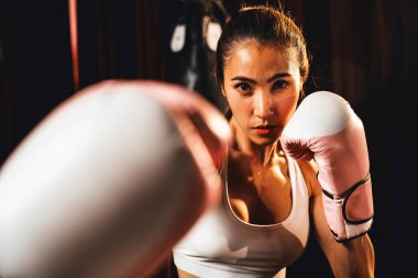 Asya 'lı kadın Muay Thai boksörü kamera önünde boksör yumruğu. Arka planda boks ekipmanıyla spor salonunda poz vermeye hazır. Kararlı gözler ve meydan okumaya hazır olun. Impetus