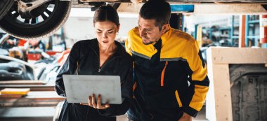 Kaldırılmış arabanın altında birlikte çalışan iki araç tamircisi, dizüstü bilgisayarla araba teftişi yapıyor. Üniformalı otomotiv teknisyeni dikkatle tanısal sorun giderme yapıyor. Panorama Oxus