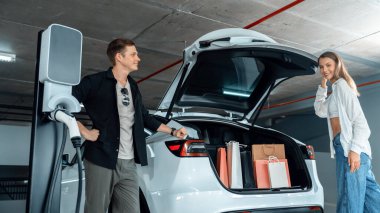 Genç çift EV elektrikli arabasıyla alışveriş merkezinin otoparkına gidiyor şehir merkezindeki şarj oluyor yeşil temiz şarj edilebilir elektrikli araç iç organları ile kentsel sürdürülebilirlik yaşam tarzını gösteriyor