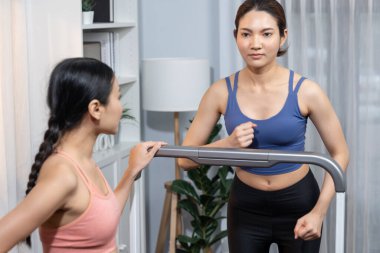 Enerjik ve güçlü atletik Asyalı kadın evde spor arkadaşı ya da antrenörü ile birlikte eliptik koşu makinesiyle koşuyor. Formda fiziğin takibi ve sağlıklı yaşam tarzına bağlılık. Kuvvetli