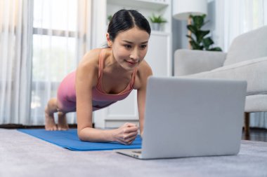 Çevrimiçi eğitim videosundaki egzersiz talimatlarını takip ederken, Asyalı genç bir kadın oturma odasının zemininde planlar yapıyor. Evde sağlıklı bir yaşam tarzı egzersizi. Denge ve dayanıklılık kavramı. Kuvvetli