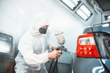 Tam korumalı ekipmanlı otomotiv işçisi sprey tabanca ya da oda atölyesindeki solunum cihazı boyasıyla araba kaportasına ustalıkla renk boyası uygular. Tamir için araba boyama servisi. Oxus.