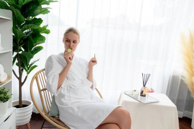 Elinde taze salatalık dilimleri tutan ve bornoz giyen bir kadın tatil köyünde ya da otelde lüks yüz derisi kaplıcalarının tadını çıkarıyor. Yüz ve güzellik tedavisi için cilt bakımı. Sessiz.