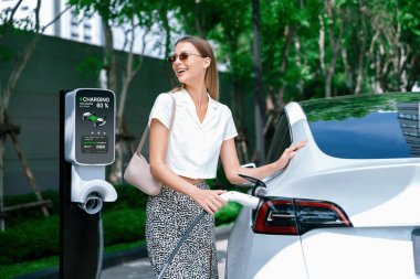 Genç bir kadın yazın yeşil sürdürülebilir şehir bahçesinde EV elektrikli arabasıyla seyahat ediyor. Kentsel sürdürülebilirlik yaşam tarzı yeşil temiz yeniden şarj edilebilir enerji elektrikli BEV araç iç organları