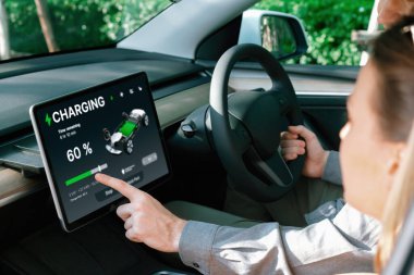 Elektrikli araba sürücüsü, arabadaki uygulama ekranında şarj durumu, menzil ve şarj limitini kontrol ediyor. Akıllı teknoloji aygıtı, EV arabasının araba aküsündeki elektrik depolama verilerini şarj ettiğini gösteriyor..