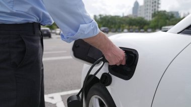 İlerici işadamı, şehir merkezinde dolaşmadan önce şarj cihazından elektrikli arabasına şarj prizi takıyor. Çevre dostu şarj edilebilir araba, sürdürülebilir ve temiz enerjiyle çalışıyor..