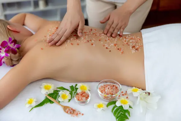 Woman customer having exfoliation treatment in luxury spa salon with warmth candle light ambient. Salt scrub beauty treatment in health spa body scrub. Quiescent