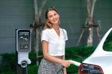 Genç bir kadın yazın yeşil sürdürülebilir şehir bahçesinde EV elektrikli arabasıyla seyahat ediyor. Kentsel sürdürülebilirlik yaşam tarzı yeşil temiz yeniden şarj edilebilir enerji elektrikli BEV araç iç organları