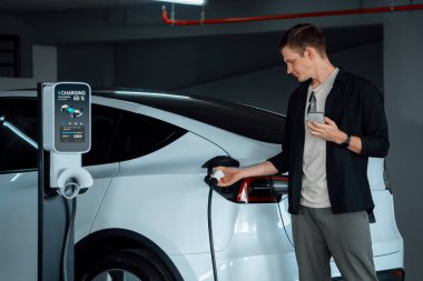 Genç adam EV elektrikli arabasıyla alışveriş merkezinin otoparkına gidiyor şehir merkezindeki şarj oluyor yeşil, temiz, yeniden şarj edilebilir elektrikli araç iç organları ile kentsel sürdürülebilirlik yaşam tarzını gösteriyor