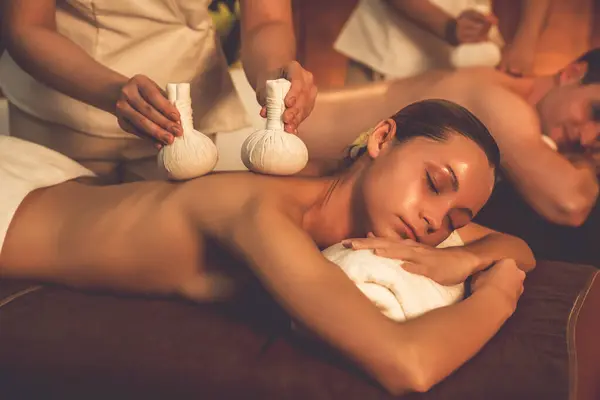 Hot herbal ball spa massage body treatment, masseur gently compresses herb bag on couple customer body. Serenity of aromatherapy recreation in warm lighting of candles at spa salon. Quiescent