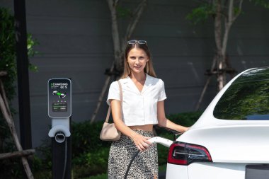Genç bir kadın yazın yeşil sürdürülebilir şehir bahçesinde EV elektrikli arabasıyla seyahat ediyor. Kentsel sürdürülebilirlik yaşam tarzı yeşil temiz yeniden şarj edilebilir enerji elektrikli BEV araç iç organları
