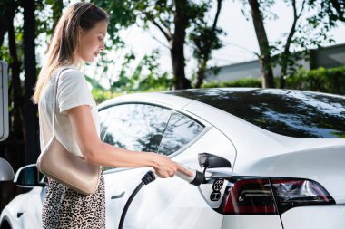 Genç bir kadın yazın yeşil sürdürülebilir şehir bahçesinde EV elektrikli arabasıyla seyahat ediyor. Kentsel sürdürülebilirlik yaşam tarzı yeşil temiz yeniden şarj edilebilir enerji elektrikli BEV araç iç organları
