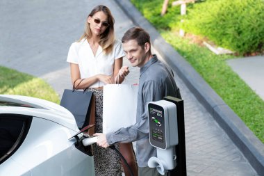 Genç çift yazın yeşil sürdürülebilir şehir bahçesinde EV elektrikli araba ile seyahat ederken, yeşil temiz yeniden şarj edilebilir elektrikli araç iç organları ile kentsel sürdürülebilirlik yaşam tarzını gösteriyor.