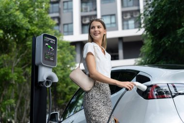 Genç bir kadın yazın yeşil sürdürülebilir şehir bahçesinde EV elektrikli arabasıyla seyahat ediyor. Kentsel sürdürülebilirlik yaşam tarzı yeşil temiz yeniden şarj edilebilir enerji elektrikli BEV araç iç organları
