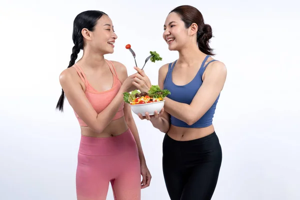 Two young sporty Asian women in sportswear holding salad bowl fill with fruit and vegetable. Natural youthful and fit body lifestyle people with balance nutrition on isolated background. Vigorous