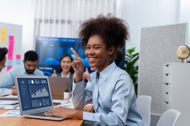 Bir grup ofis çalışanı ile birlikte arka planda ekranda gösterim paneliyle buluşan mutlu Afrikalı genç bir iş kadını portresi. Takım toplantısında kendine güveni tam bir ofis hanımı. Uyum