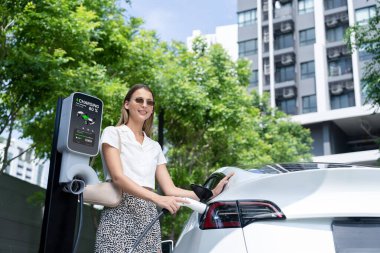Genç bir kadın yazın yeşil sürdürülebilir şehir bahçesinde EV elektrikli arabasıyla seyahat ediyor. Kentsel sürdürülebilirlik yaşam tarzı yeşil temiz yeniden şarj edilebilir enerji elektrikli BEV araç iç organları