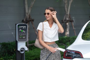 Genç bir kadın yazın yeşil sürdürülebilir şehir bahçesinde EV elektrikli arabasıyla seyahat ediyor. Kentsel sürdürülebilirlik yaşam tarzı yeşil temiz yeniden şarj edilebilir enerji elektrikli BEV araç iç organları