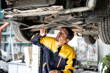 Araç tamircisi kaldırılmış aracın altından araba teftişi yapıyor. Üniformalı otomotiv teknisyeni araba akslarını ve şasi parçalarını dikkatle teşhis ediyor ve kontrol ediyor. Oxus.
