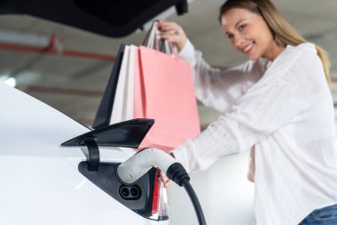 Genç bir kadın EV elektrikli arabasıyla alışveriş merkezinin otoparkına gidiyor şehir merkezinde elektrik iç organlarının yeşil temiz şarj edilebilir enerji ile sürdürülebilirlik yaşam tarzını gösteriyor.