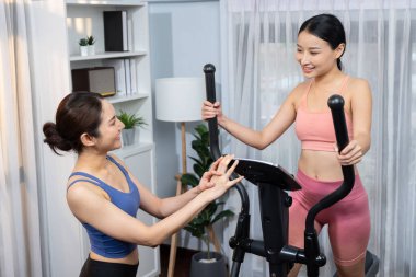 Enerjik ve güçlü atletik Asyalı kadın evde spor arkadaşı ya da antrenörü ile birlikte eliptik koşu makinesiyle koşuyor. Formda fiziğin takibi ve sağlıklı yaşam tarzına bağlılık. Kuvvetli