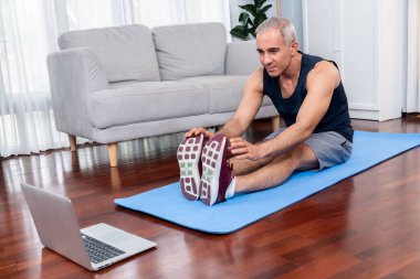 Aktif ve formda bir adam, evde egzersiz yapmadan önce ısınma ve esneme hareketleri yapıyor ve çevrimiçi fitness videosu izliyor. Emekliler için sağlıklı yaşam tarzı konsepti. Etki