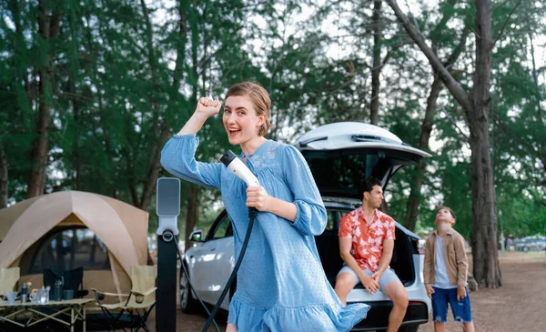Outdoor adventure and family vacation camping at sea travel by eco friendly car. Cheerful woman or mother holding, pointing EV charger point with playful and happiness posture in campsite. Perpetual