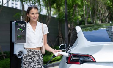 Genç bir kadın yazın yeşil sürdürülebilir şehir bahçesinde EV elektrikli arabasıyla seyahat ediyor. Kentsel sürdürülebilirlik yaşam tarzı yeşil temiz yeniden şarj edilebilir enerji elektrikli BEV araç iç organları