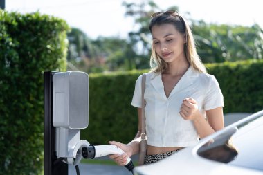 Genç bir kadın yazın yeşil sürdürülebilir şehir bahçesinde EV elektrikli arabasıyla seyahat ediyor. Kentsel sürdürülebilirlik yaşam tarzı yeşil temiz yeniden şarj edilebilir enerji elektrikli BEV araç iç organları
