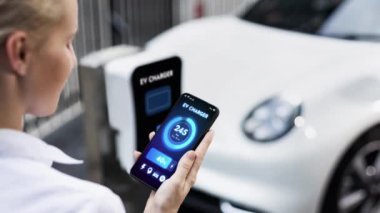 Akıllı telefon akü durumu arayüzünü elinde tutan iş kadını akıllı EV mobil uygulama ile otomobil parkındaki şarj istasyonundan elektrik şarj eden çevre dostu EV araba. İnceleyin