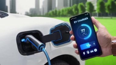 İşadamı çevre yeşil şehir parkındaki şarj istasyonundan şarj olurken akıllı telefon ekranından EV batarya durum mobil uygulamasını kontrol ediyor. Elektrikli arabanın gelecekteki enerji sürdürülebilirliği. İnceleyin