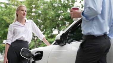 İlerici işadamı ve iş kadını, şehir merkezinde dolaşmadan önce şarj cihazını şarj istasyonundan elektrikli arabaya takıyor. Çevre dostu şarj edilebilir araba temiz enerjiyle çalışıyor..
