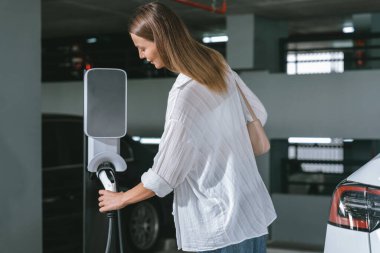 Genç bir kadın EV elektrikli arabasıyla alışveriş merkezinin otoparkına gidiyor şehir merkezinde elektrik iç organlarının yeşil temiz şarj edilebilir enerji ile sürdürülebilirlik yaşam tarzını gösteriyor.
