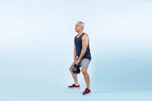 Full body length shot athletic and sporty senior man doing squat with kettlebell for body workout on isolated background. Healthy active physique and body care lifestyle after retirement. Clout