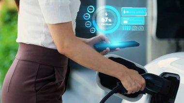 Modern yaşam tarzı kadın elektrikli arabasına EV şarj aleti yerleştiriyor ve akıllı telefondan EV batarya durumunu kontrol ediyor. Gelecekçi enerji sürdürülebilirliği ve elektrikli araba. İnceleyin