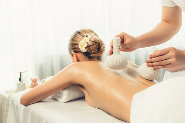 Hot herbal ball spa massage body treatment, masseur gently compresses herb bag on woman body. Tranquil and serenity of aromatherapy recreation in day lighting ambient at spa salon. Quiescent