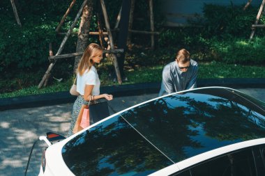 Genç çift yazın yeşil sürdürülebilir şehir bahçesinde EV elektrikli araba ile seyahat ederken, yeşil temiz yeniden şarj edilebilir elektrikli araç iç organları ile kentsel sürdürülebilirlik yaşam tarzını gösteriyor.