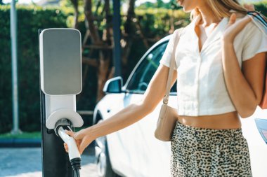 Genç bir kadın yazın yeşil sürdürülebilir şehir bahçesinde EV elektrikli arabasıyla seyahat ediyor. Kentsel sürdürülebilirlik yaşam tarzı yeşil temiz yeniden şarj edilebilir enerji elektrikli BEV araç iç organları