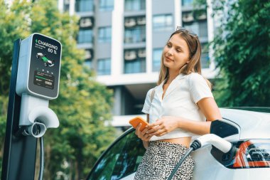Genç bir kadın yazın yeşil sürdürülebilir şehir bahçesinde EV elektrikli arabasıyla seyahat ediyor. Kentsel sürdürülebilirlik yaşam tarzı yeşil temiz yeniden şarj edilebilir enerji elektrikli BEV araç iç organları