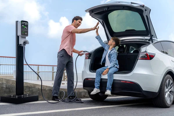 Family road trip vacation traveling by the sea with electric car, father and son high five after reach destination at EV charging station by the seashore. Eco-friendly car for environment. Perpetual