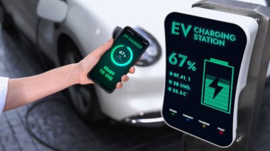 Akıllı telefon ekran bataryasını elinde tutan iş adamı akıllı EV mobil uygulama ile otoparktaki şarj istasyonundan elektrik şarj eden EV araba. İnceleyin