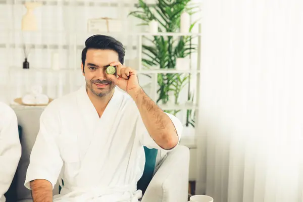 Man holding slices of fresh cucumber and wearing bathrobe enjoying luxurious facial skincare spa in resort or hotel. Skin treatment for face and beauty care. Quiescent