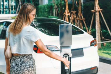 Genç bir kadın yazın yeşil sürdürülebilir şehir bahçesinde EV elektrikli arabasıyla seyahat ediyor. Kentsel sürdürülebilirlik yaşam tarzı yeşil temiz yeniden şarj edilebilir enerji elektrikli BEV araç iç organları