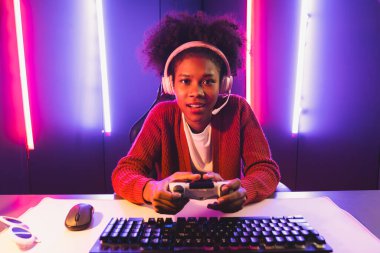 Oyun flamasının sunucu kanalı, joystick ile online oyun oynayan Afrikalı kız, mikrofondan izleyici medyasıyla konuşuyor. Esport yetenekli takım oyuncuları neon renkli aydınlatma odasında. Tatlandırıcı.