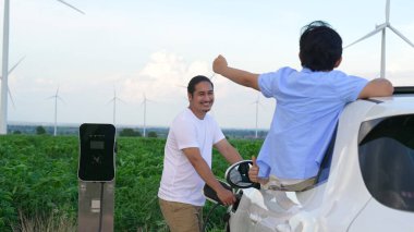 İlerici mutlu aile kavramı elektrikli araçla rüzgar çiftliğinde eğleniyor. Rüzgar türbini jeneratöründen gelen temiz yenilenebilir enerji ile şarj edilen elektrikli araç..