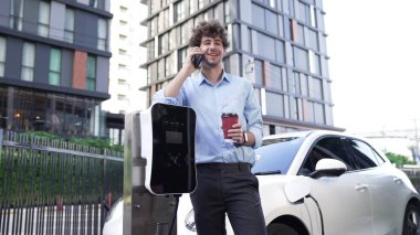 Takım elbiseli, hırslı bir işadamı elektrik kablosu ve yenilenebilir enerji enerjili bir elektrikli araçla şarj istasyonunda dururken elektrikli aracına yaslanıyor..
